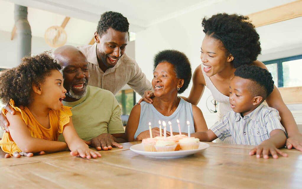 An elderly couple celebrates the birth of the older woman as her child, son-in-law, and grandchildren laugh and enjoy the day
