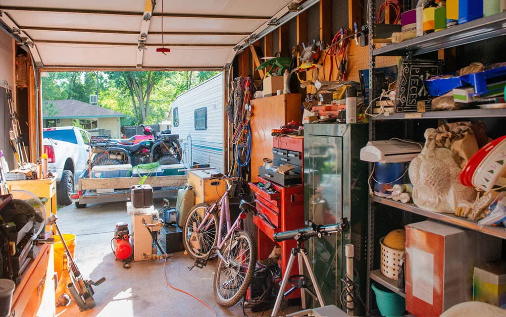 View from inside a home's garage that is filled with tools, boxes, and other household items as the driveway is filled with cars, motorcycles, and an RV