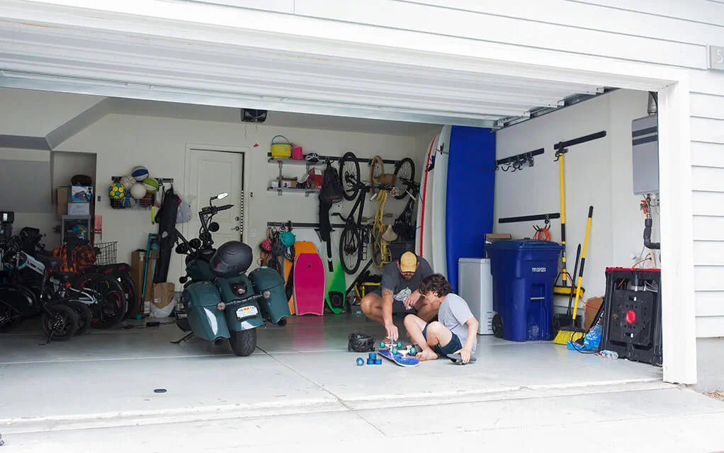A father and soon work on fixing a skateboard in their garage as a motorcycle sits idle next to them, with other home goods in the garage