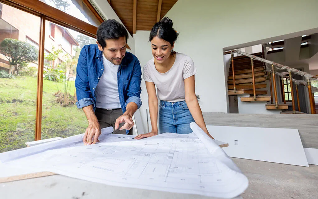 A young couple stands over a table and views the blueprint layout of their home that is undergoing a renovation