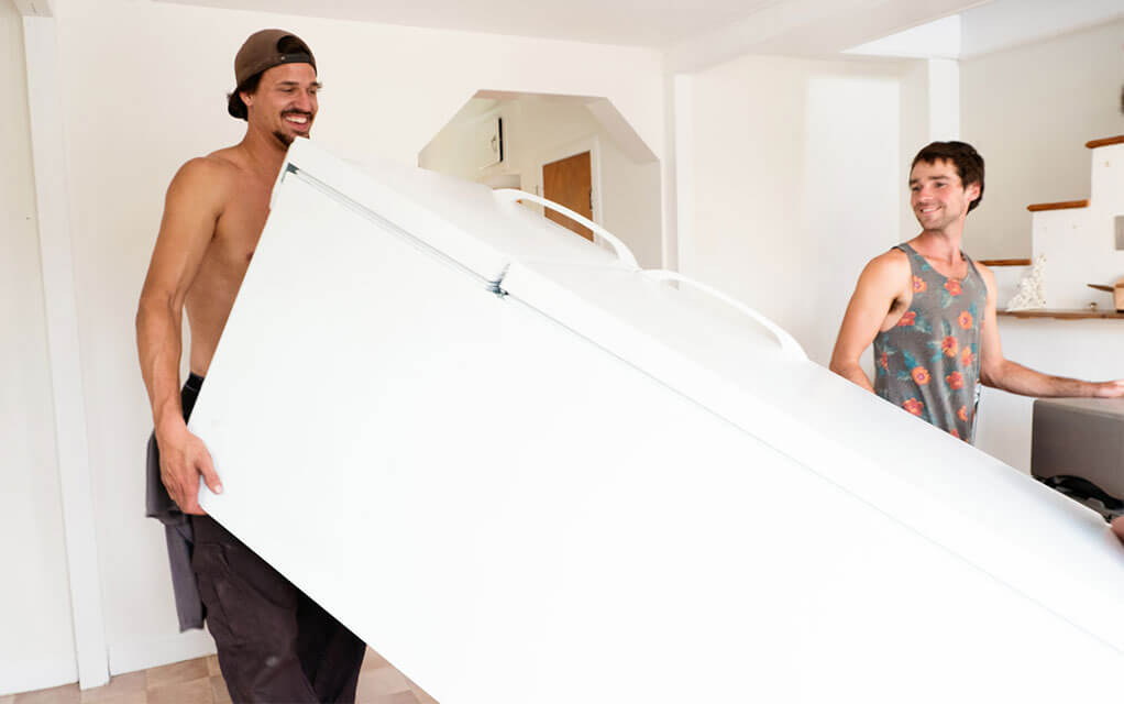 A man holds a white refrigerator at an angle as another man spots him as they move the fridge out of the house