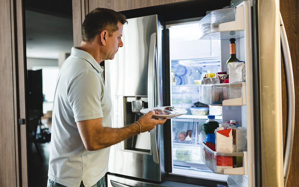 A man places a plate of covered food into his home's refrigerator, as food sits inside of it