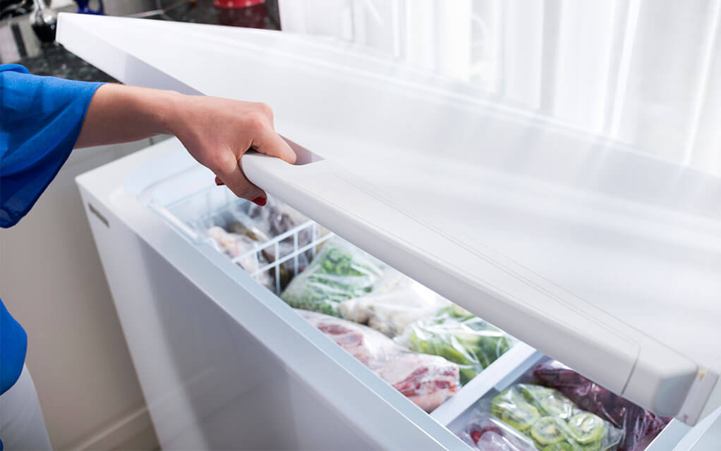 A person opens their home's chest freezer, revealing a number of frozen food items inside