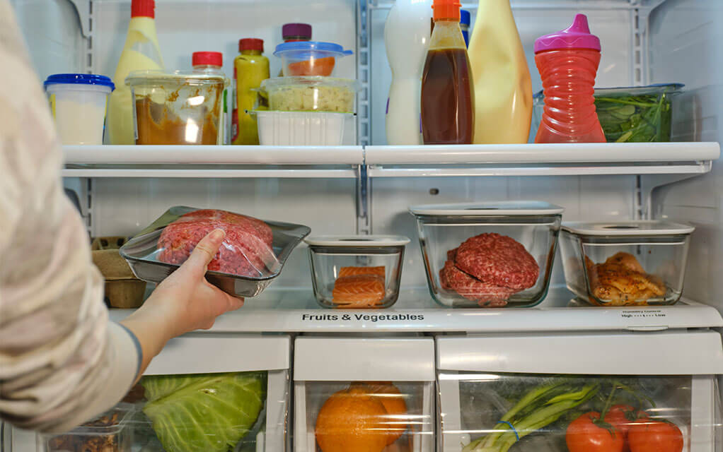 View of an open fridge filled with food, such as meat, fish, vegetables, milk, and other foodstuffs, as a person places some meat on the fridge counter
