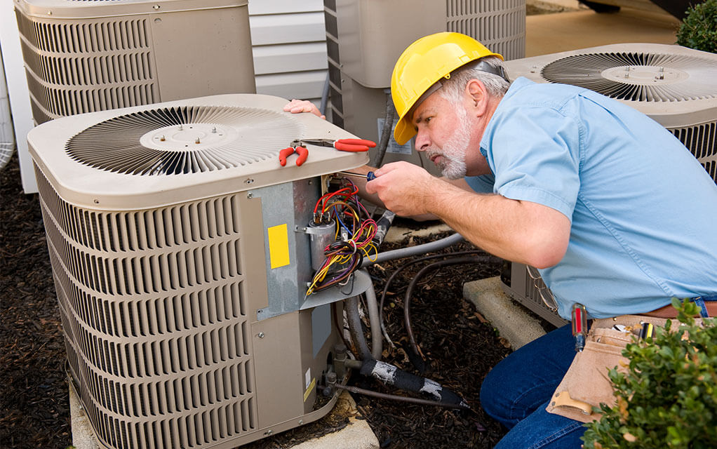 An HVAC technician with a hardhat on has the maintenance panel of an outdoor HVAC unit off as he examines and manipulates the wiring of the unit  