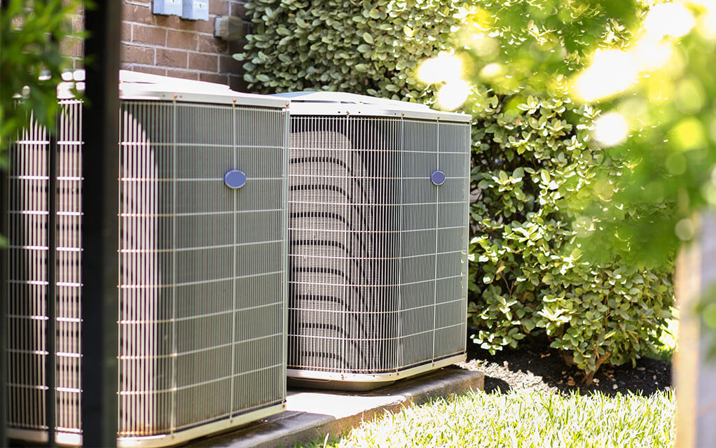 Two outdoor HVAC units sit outside a home next to each other on a bright and clear day