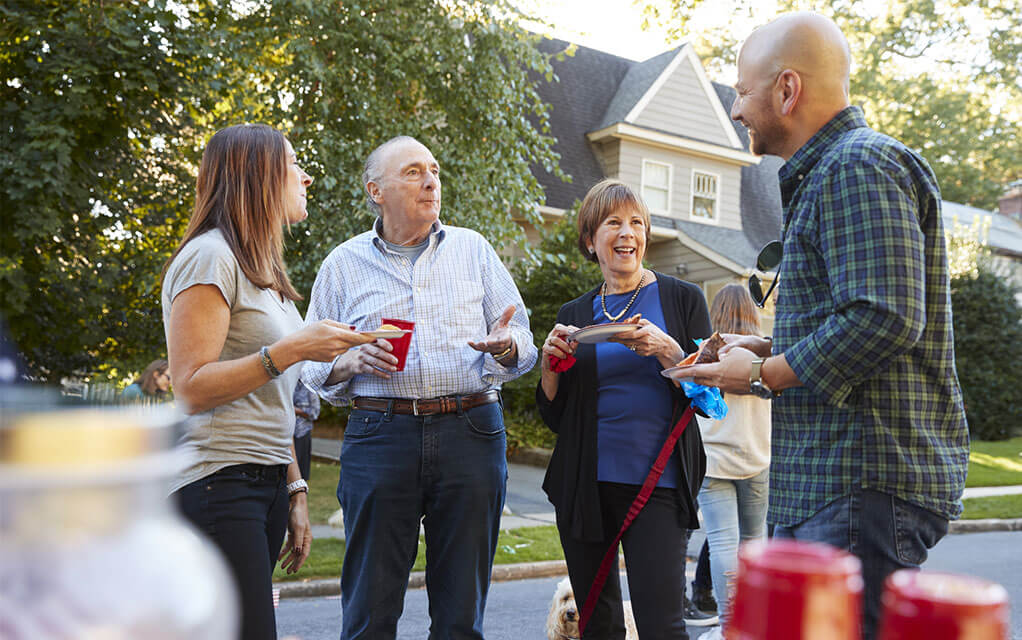 An older couple holding red drink cups talk pleasantly with a younger couple in their neighborhood street during a party