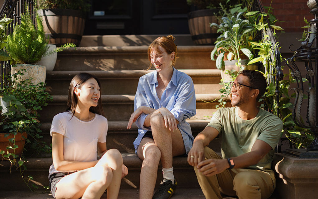 Three young people - two women and one man - sit on the steps leading up to a town house and smile and laugh as they talk with each other