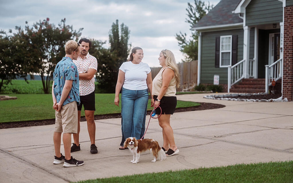 Two couples have a conversation with each other in one of the couples' driveway on a cloudy afternoon as a one of the women holds a dog's lesh