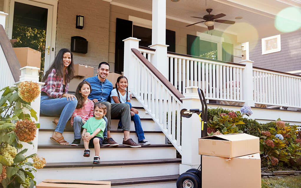 A happy family sits on the steps of their front porch in front of their new home, smiling as moving boxes sit behind them and on a moving dolly at the foot of the stairs