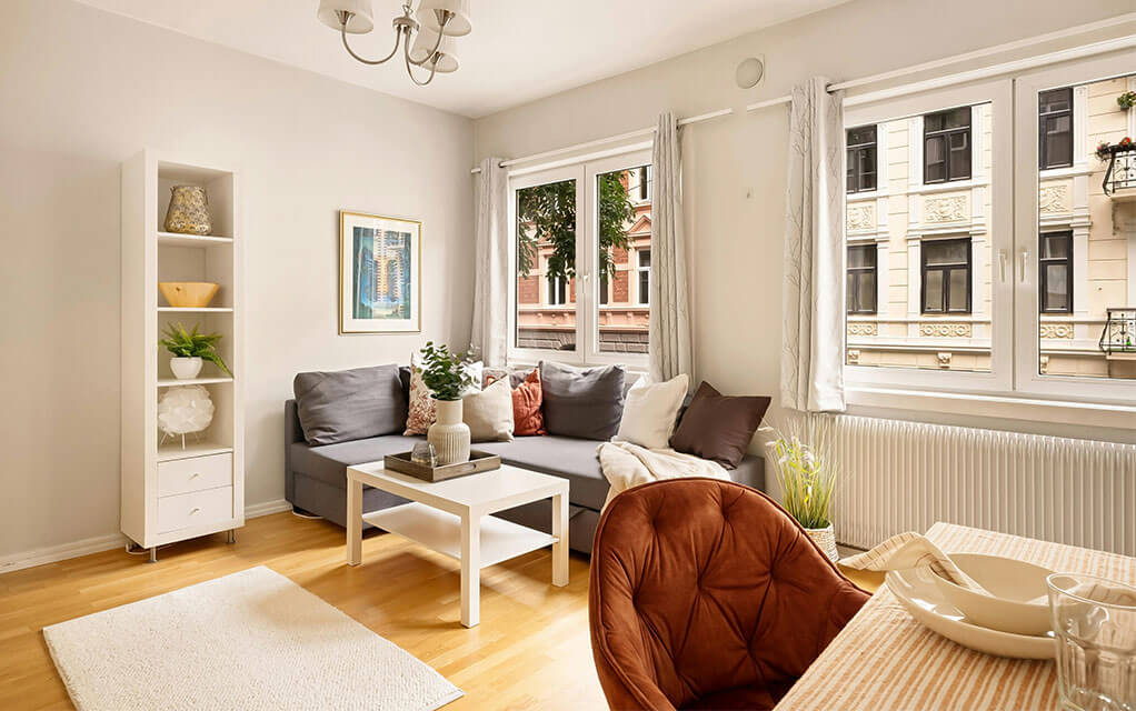 View of an upscale interior apartment living room, with an L shaped couch in the corner and decor on shelving and tables as the windows let in bright light