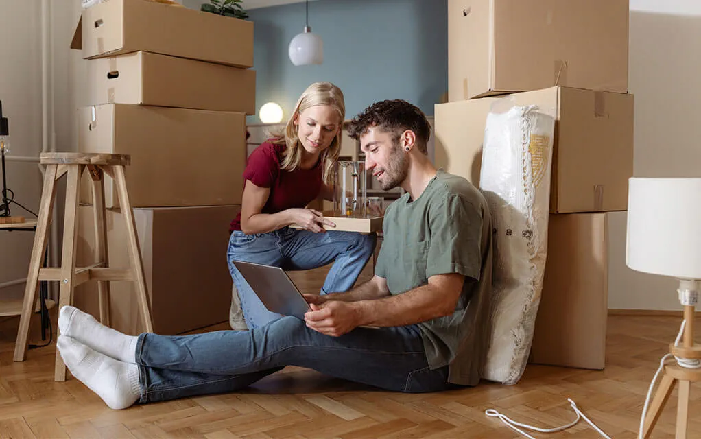 A young couple looks at something on the man's laptop with two columns of moving boxes on either side of them both