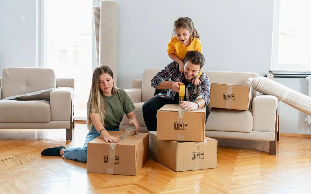 A young family tape up moving boxes as their young daughter stands over her dad's shoulders as he tapes up a box