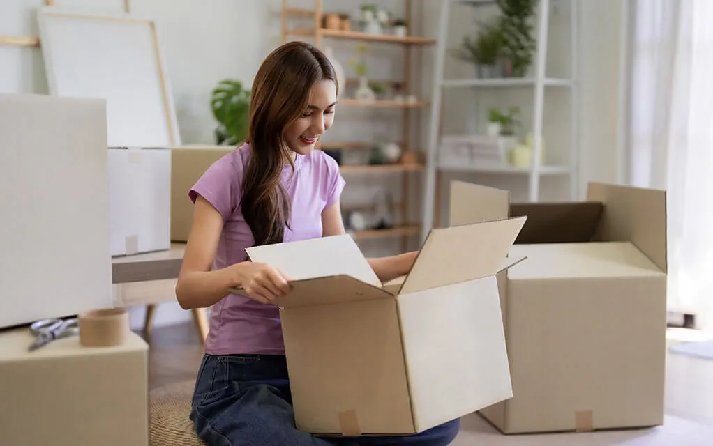 A young woman smiles as she has an open moving box in her lap with other moving boxes, both open and unopen, lay around her in her living room