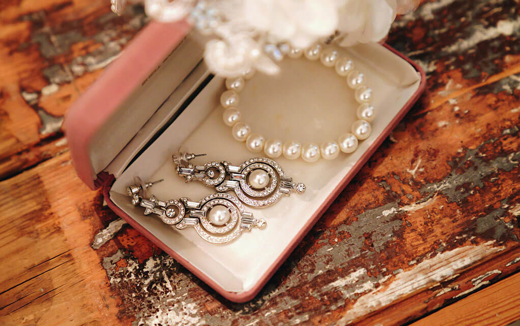 Fancy diamond earrings and a pearl bracelet sit in an open jewelry box on an old table