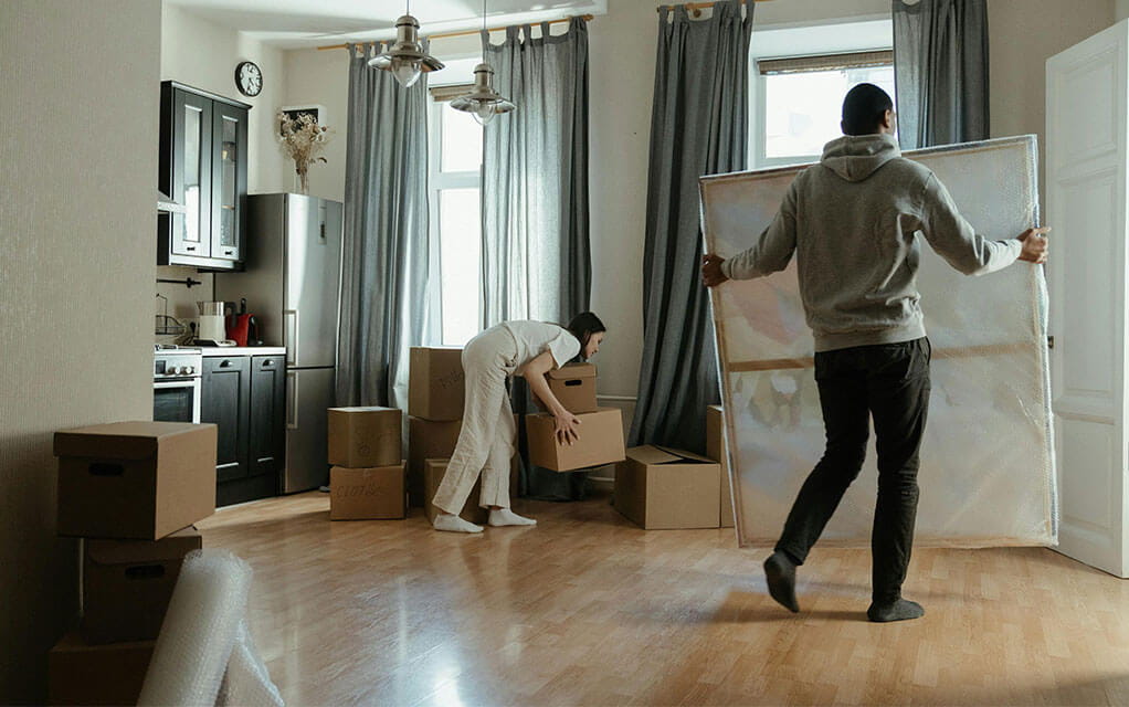 A couple are packing stuff away for a move in their home, as the woman stacks boxes and the man carries a piece of protected artwork