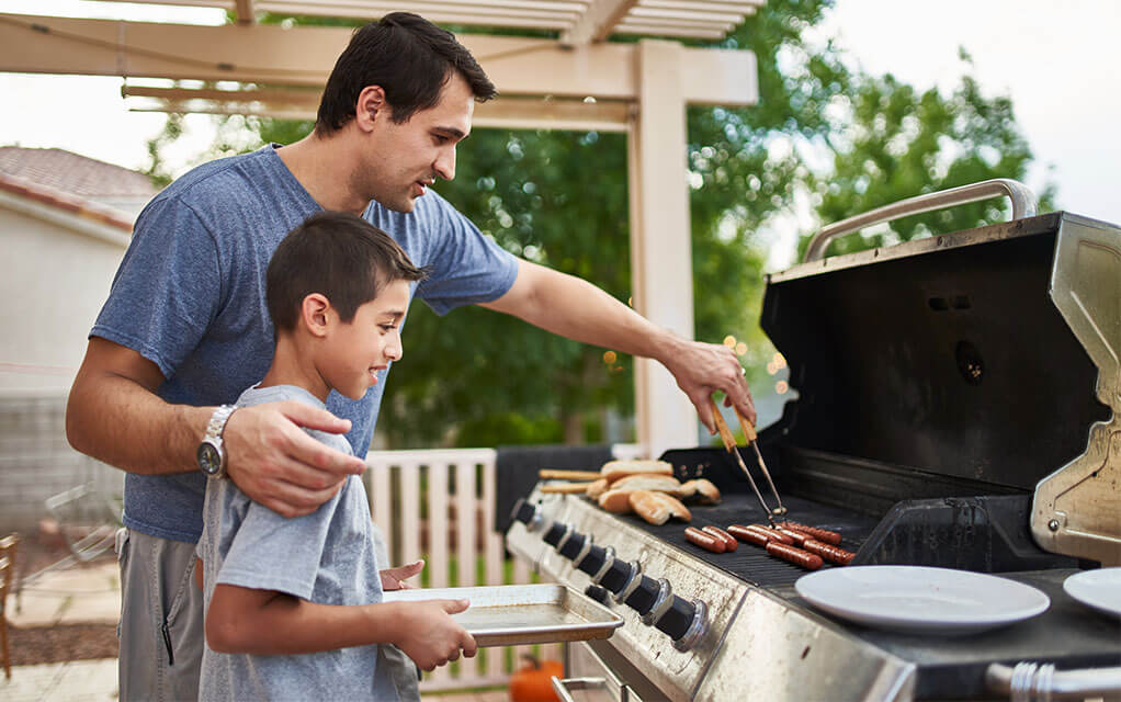 A man is helping get his young son the hot dogs off the grill and onto the pan he is holding on their back deck on a cloudy day