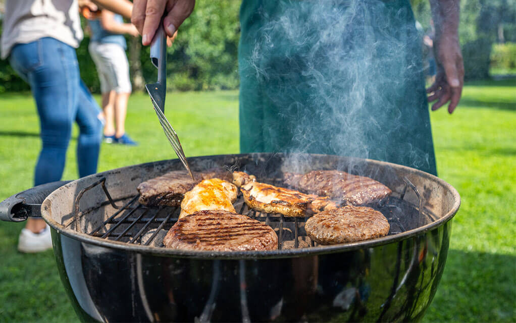A man attends to and flips some chicken and burgers on his charcoal grill and his wife plays with their child in the background in their backyard on a sunny and clear day