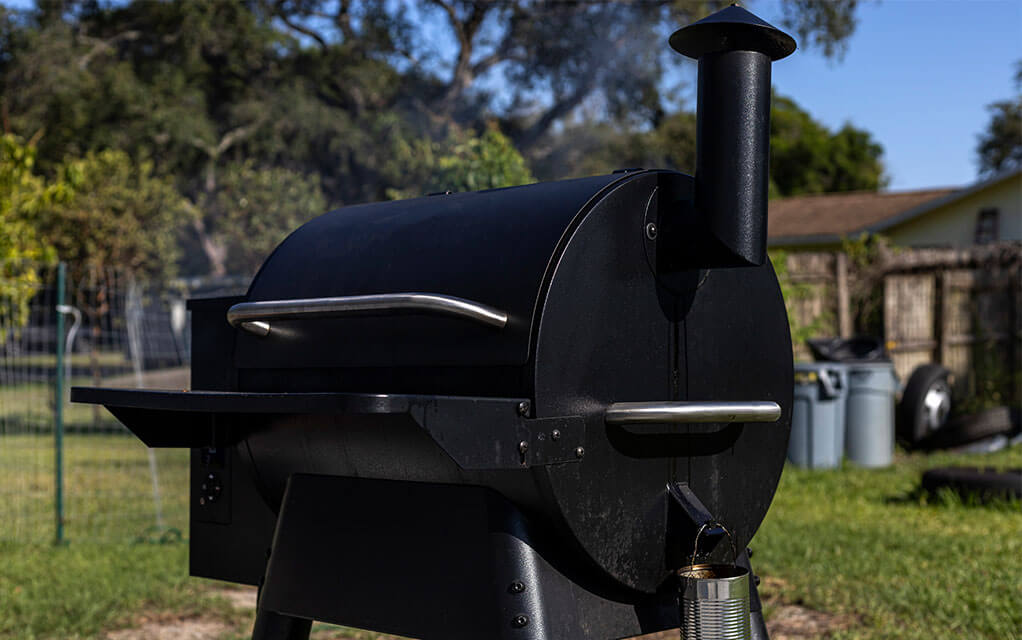 A black pellet smoker grill sits in the backyard of a home on a bright and sunny day as smoke emanates from it, indicating something is cooking inside