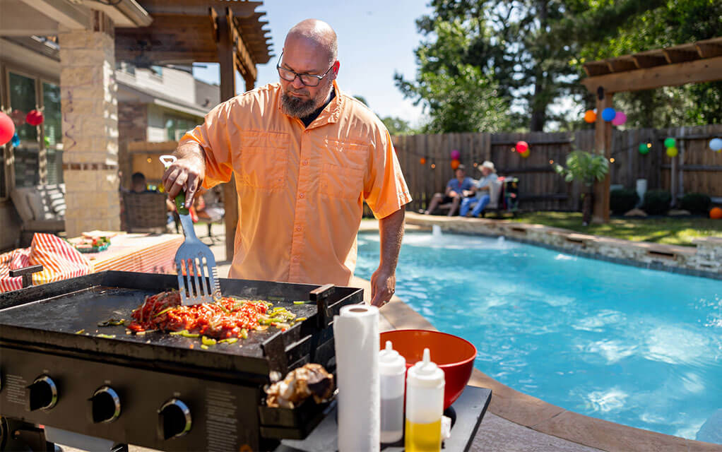 A man is cooking food on a flat top propane grill next to his in-ground pool in the backyard of his home on a bright and sunny day