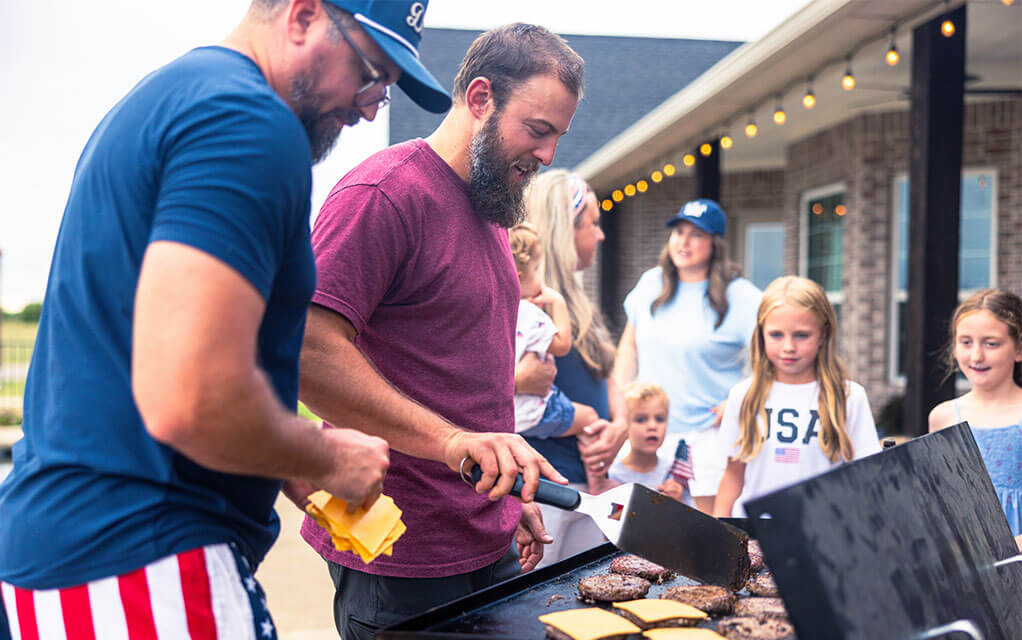 Two men work on grilling and preparing hamburgers as their wives and children are in the background talking and waiting to eat on the back porch of an upper middle class home on a clear day