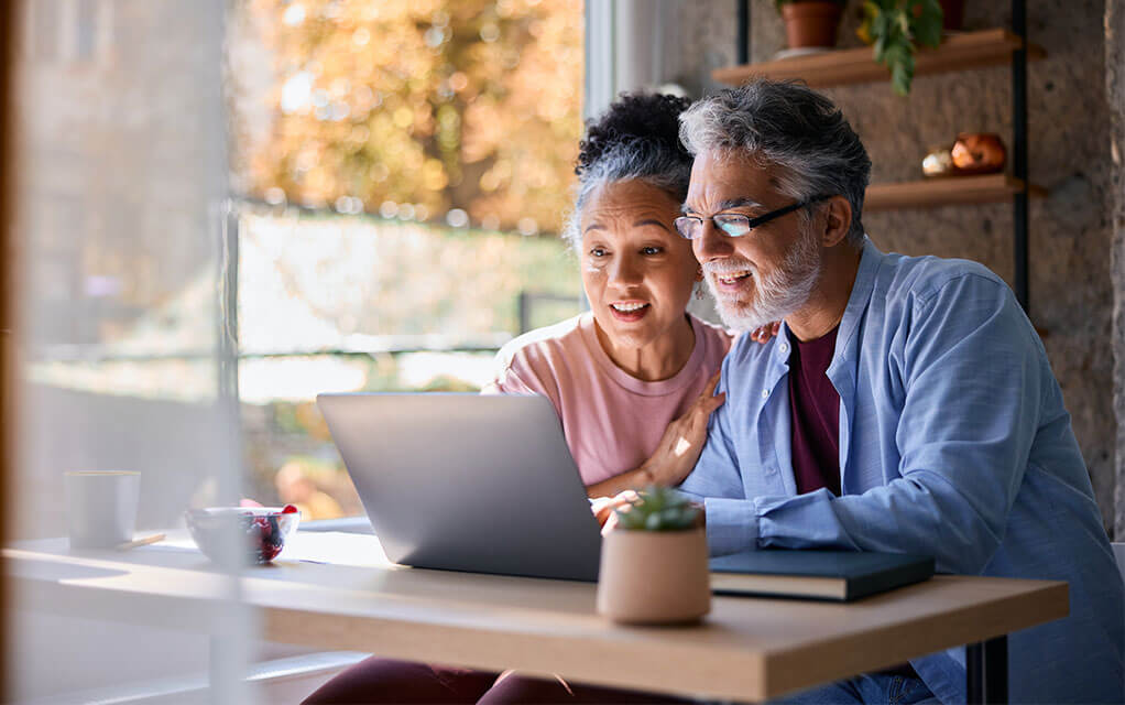 An older couple sits in their home's kitchen at the table as they look at their laptop with surprise