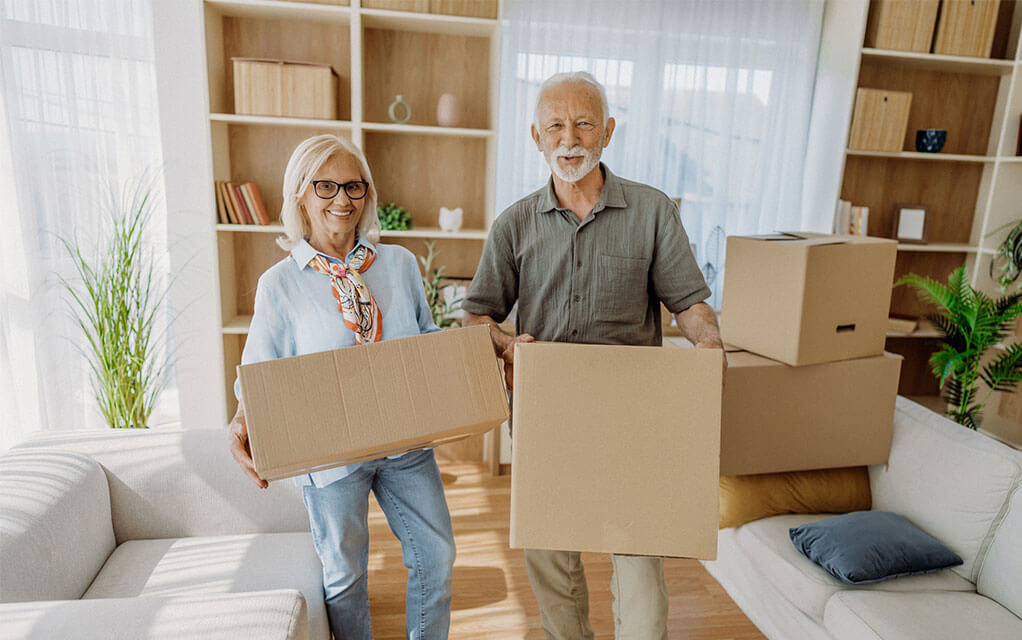 A retired couple holds moving boxes in their old home as they pack things up for their upcoming move