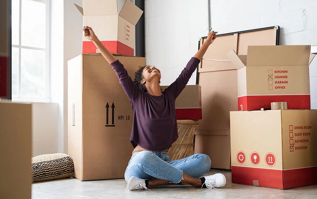 A joyful woman with her arms raised and head held high is happy surrounded by open and unopened moving boxes