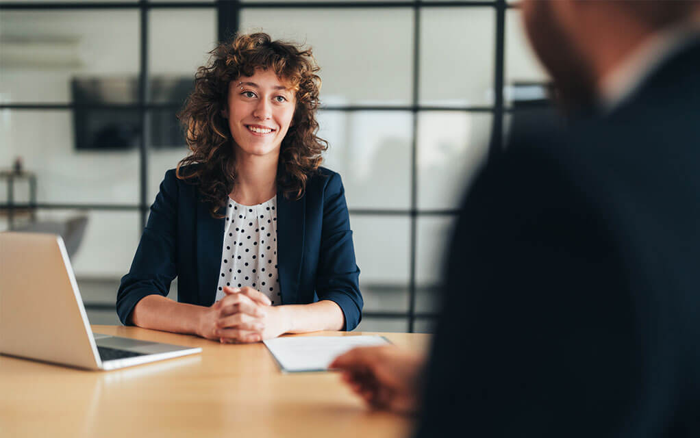 A young woman sits at a conference table in an office with an open laptop and notebook in front of her as she interviews with a man sitting across from her for a job