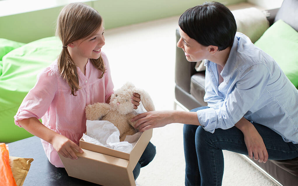 A young girl sits on the side of a table with a small cardboard box containing a teddy bear in it while her mother sits opposite from her, smiling as she holds the bear's hand