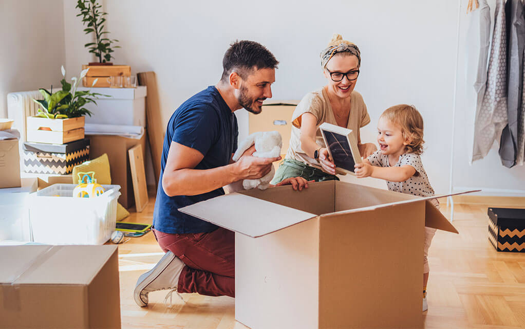 A young couple and their toddler daughter smile and laugh as they put things into a large moving box, as they are surrounded by other packed boxes and items
