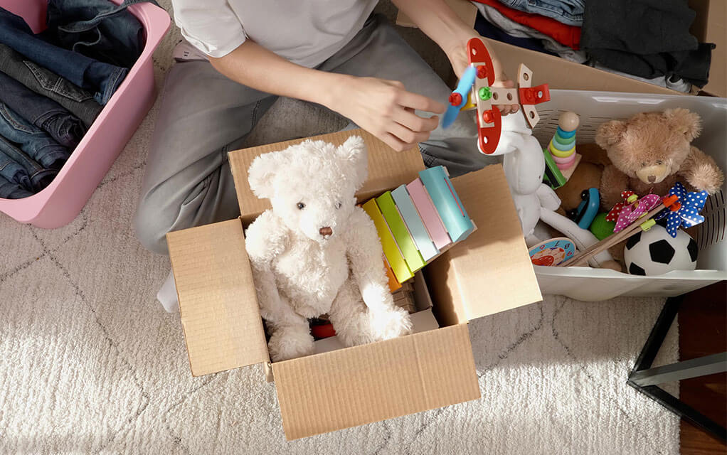 A small child is sitting on the floor and pulling toys from a clear plastic bin as a cardboard box with a teddy bear and other toys in it sits on the floor in front of him