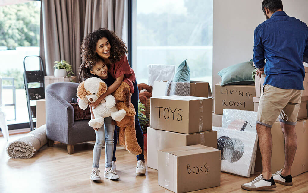 A mother gives her child a hug as they hold a large stuffed animal as they watch the father pack a box, as other marked boxes sit around them, including boxes that say TOYS and BOOKS