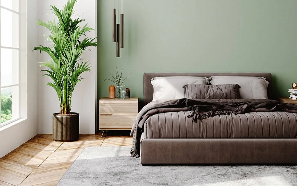 A nice bed with brown sheets and bedding sits in a nice and bright bedroom with a large plant in the corner of the room