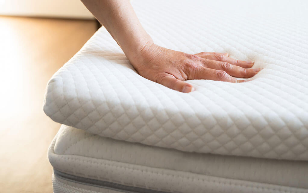 A person places their hand onto a memory foam mattress topper in their bedroom