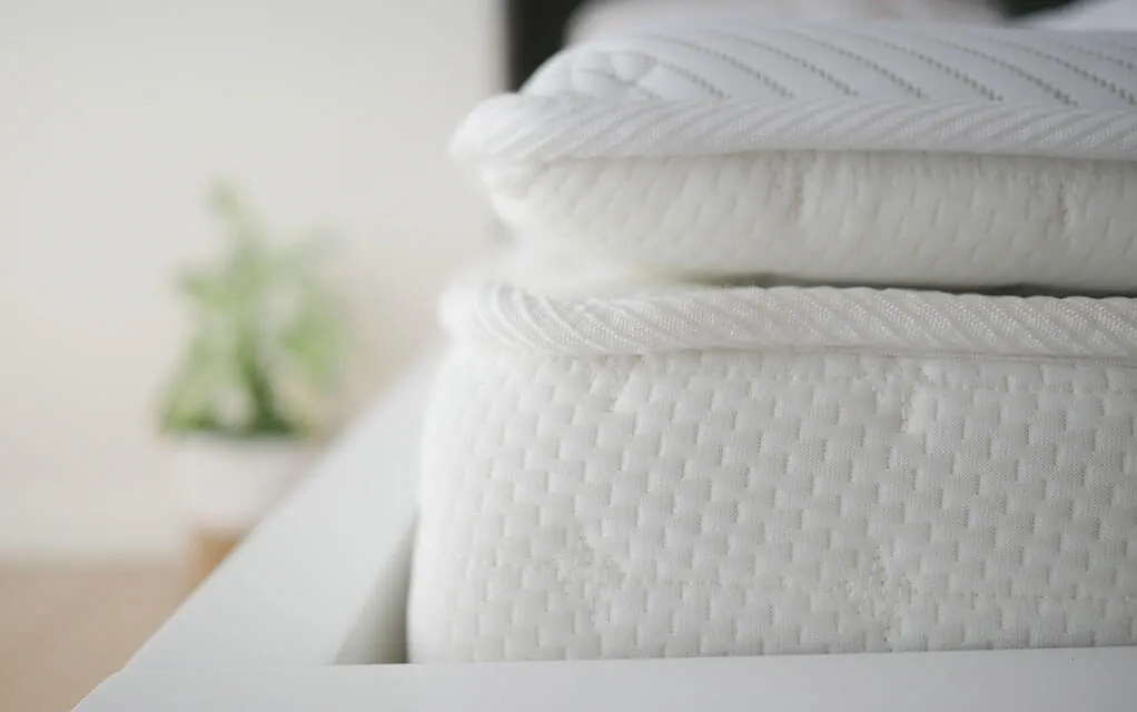View of the corner of a mattress and a mattress topper in a bedroom that is missing its covers