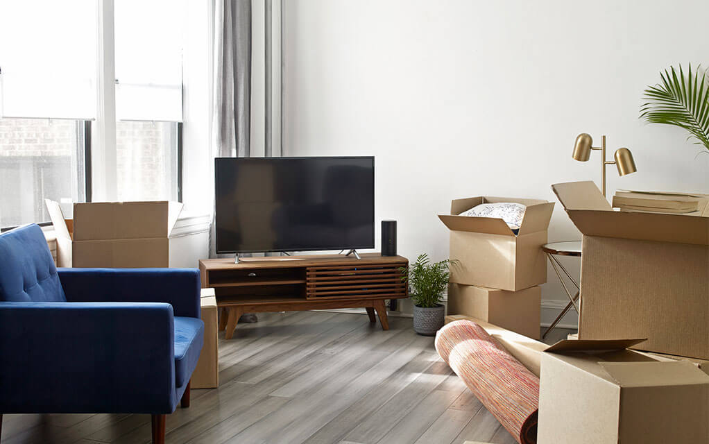 A flat screen TV sits on an entertainment center in an apartment living room, as moving boxes, both packed and unpacked lie around the rest of the room