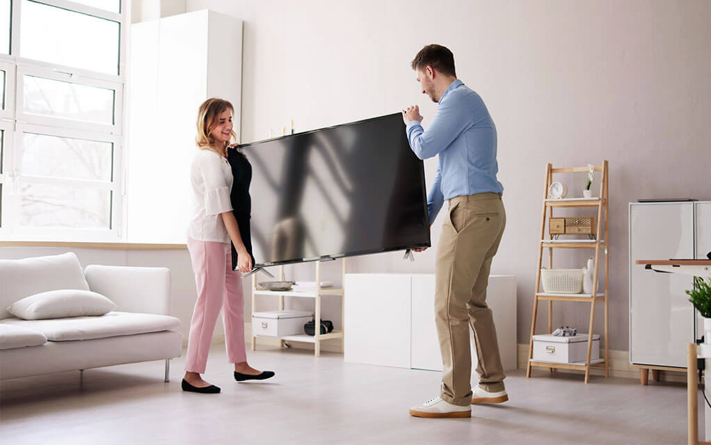 A young couple maneuvers a TV through their living room as they try to set it up in their home on a bright day