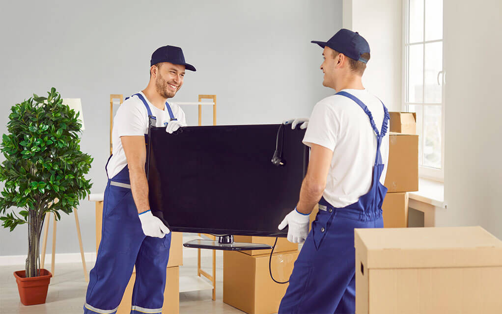 Two professional laborers handle a TV for a move inside an apartment's living room, as moving boxes are stacked around them waiting to be moved
