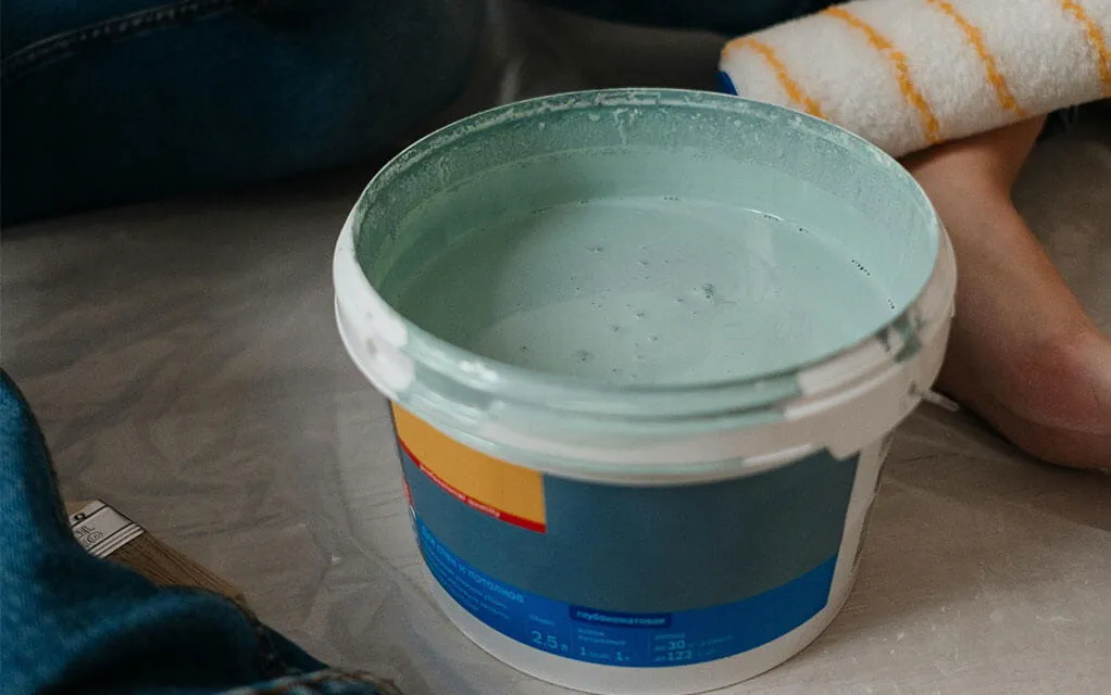 View of an open can of paint that is blue-green in color as a persons legs are visible next to it