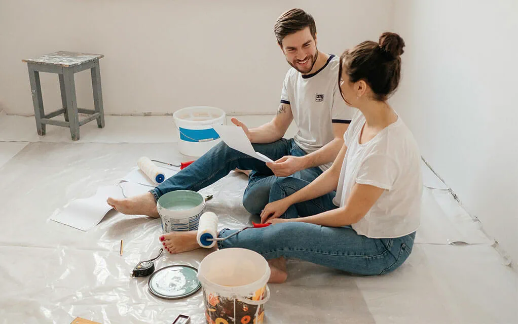 A couple sits on the floor of their prepped room smiling as open paint cans sit around them along with paint rollers