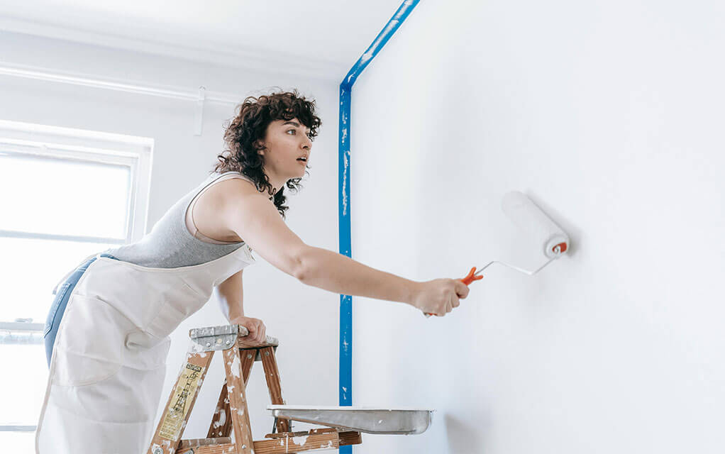 A woman stands on her paint ladder, applying a white paint to the wall with a handheld paint roller as white light shines in from outside