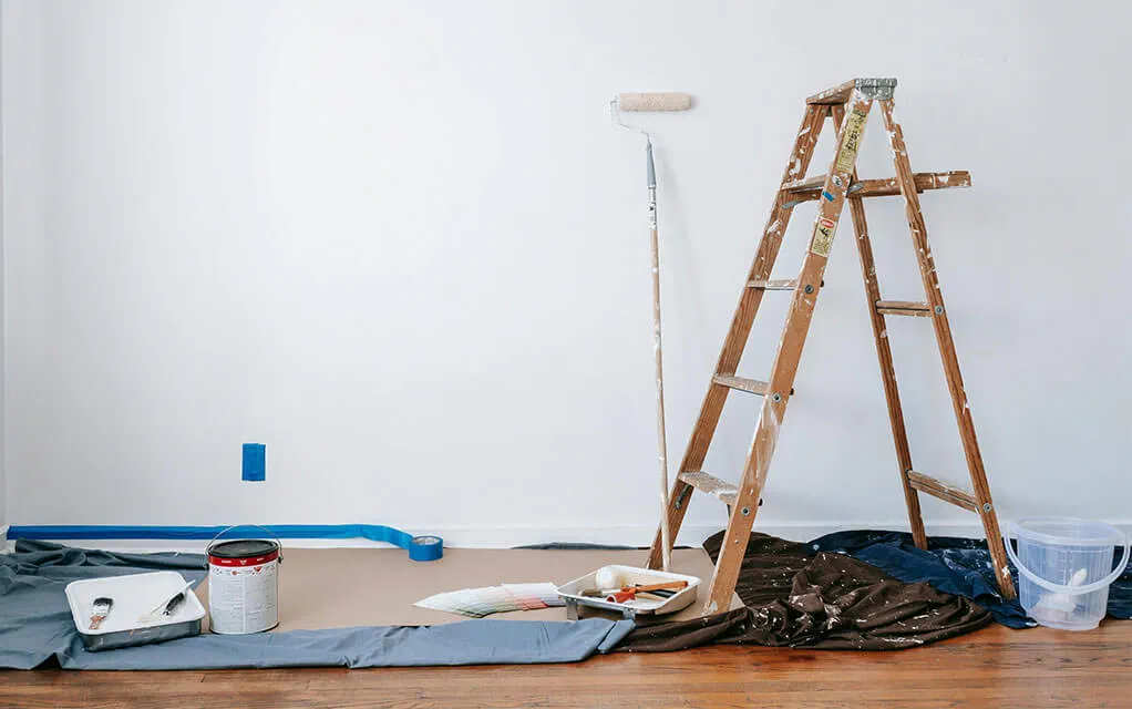 Wide view of a wall waiting to be painted as painters tap lines the wall and drop cloths lay on the floor with a ladder readied and a long paint roller awaiting paint