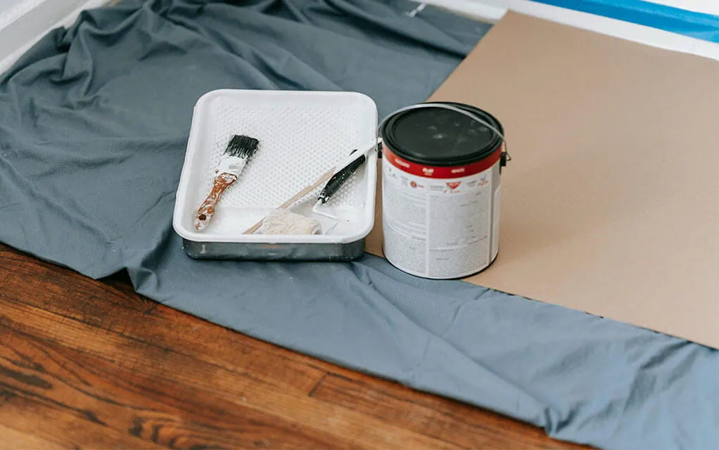 A paint can sits on top of cardboard and a drop cloth as a paint brush and scraper sit in a paint holder