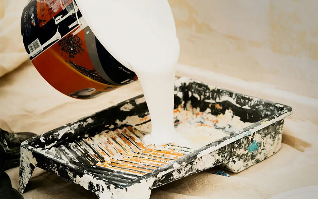 A white pain in a large paint can is being poured out into a paint holder used for a paint roller