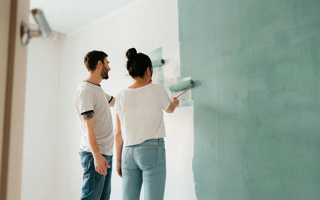 A couple is using paint rollers to apply a turquoise colored paint to the wall of a room in their home
