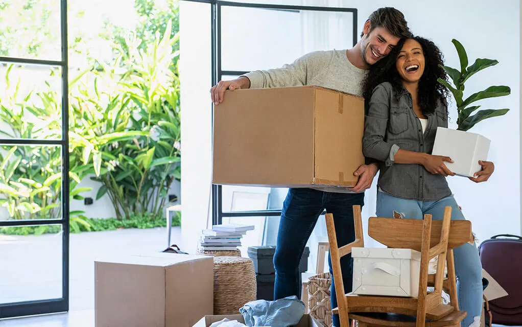 A happy couple laughs and shares a hug as the man holds a large moving box and the woman carries a smaller box, as their home is being prepared for a move