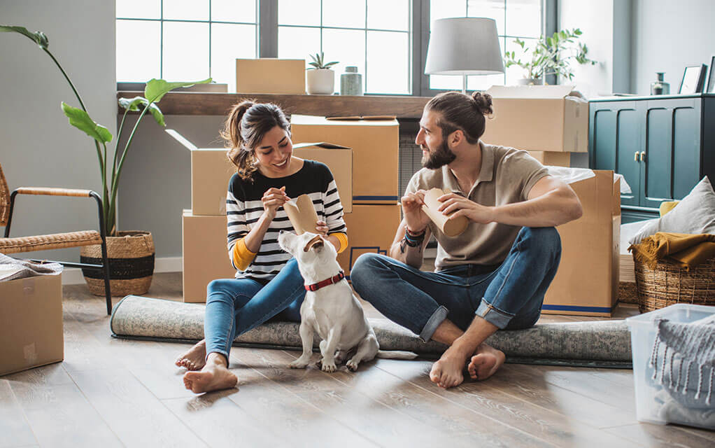 A younger couple sits on the floor of their home, with moving boxes and other household items sitting behind and around them as their dog looks at them