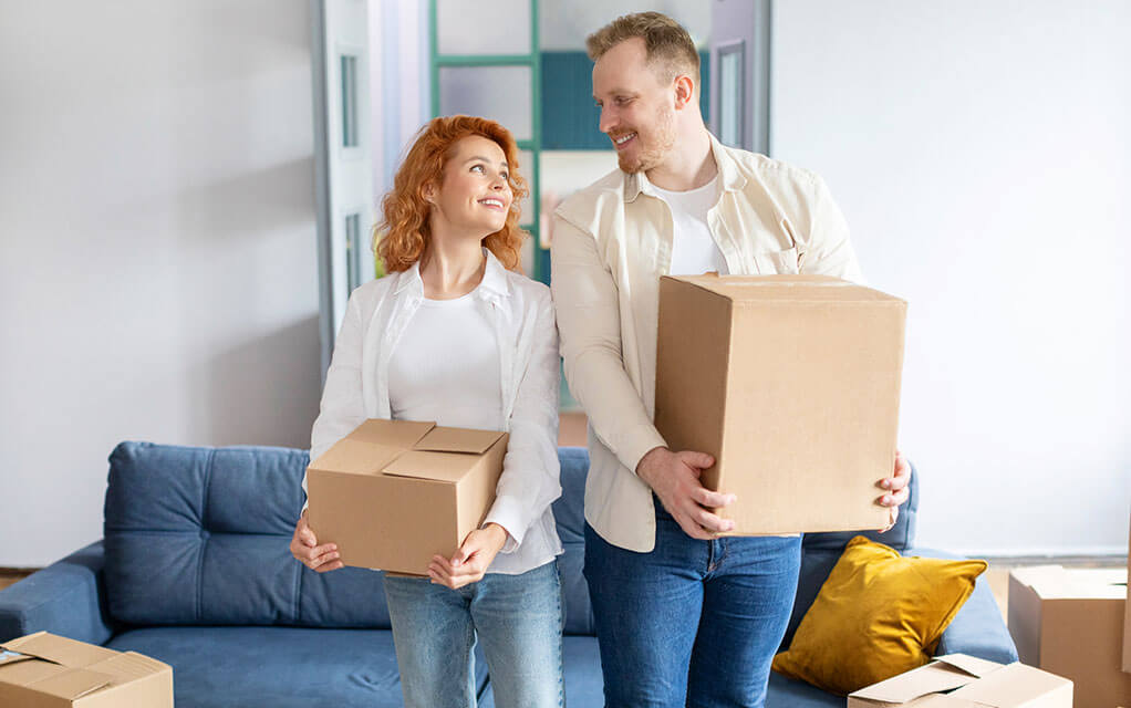 A young couple looks and smiles at each other, as they both hold packed moving boxes, as other moving boxes can be seen on the floor around them