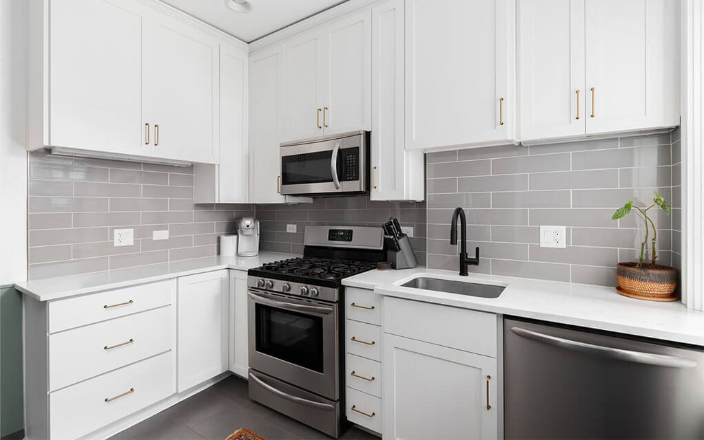 The view of a clean and immaculate kitchen, with the stainless steel oven, microwave, and dishwasher in view, along with a small sink and a few items on the counter including a small houseplant with white cabinets